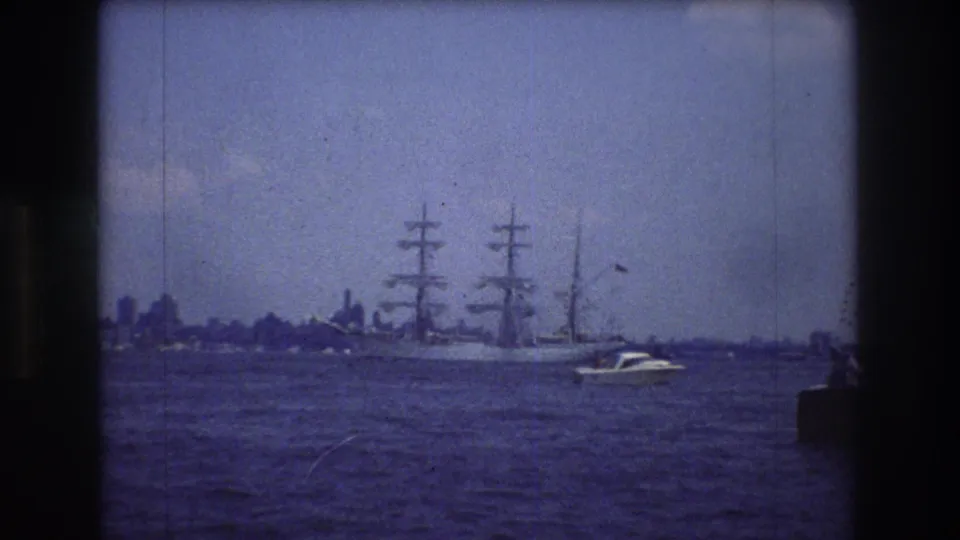 A 1976 8mm frame near New York City shows a large three-masted sailing vessel crossing open water, with a small motorboat in front and a hazy skyline behind.