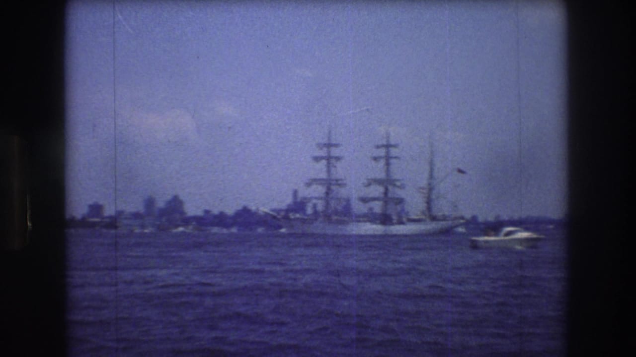 A large three-masted sailing vessel moves across open water near New York City in 1976, with a small motorboat at right and a hazy urban skyline in the distance.
