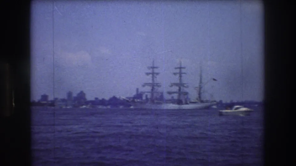 A large three-masted sailing vessel moves across open water near New York City in 1976, with a small motorboat at right and a hazy urban skyline in the distance.
