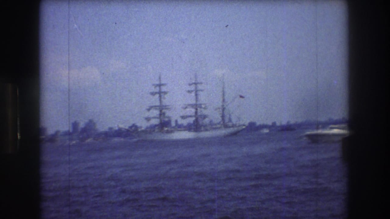 A 1976 8mm view of a multi-masted sailing ship on New York Harbor, seen across choppy water with a distant shoreline and dark frame edges.