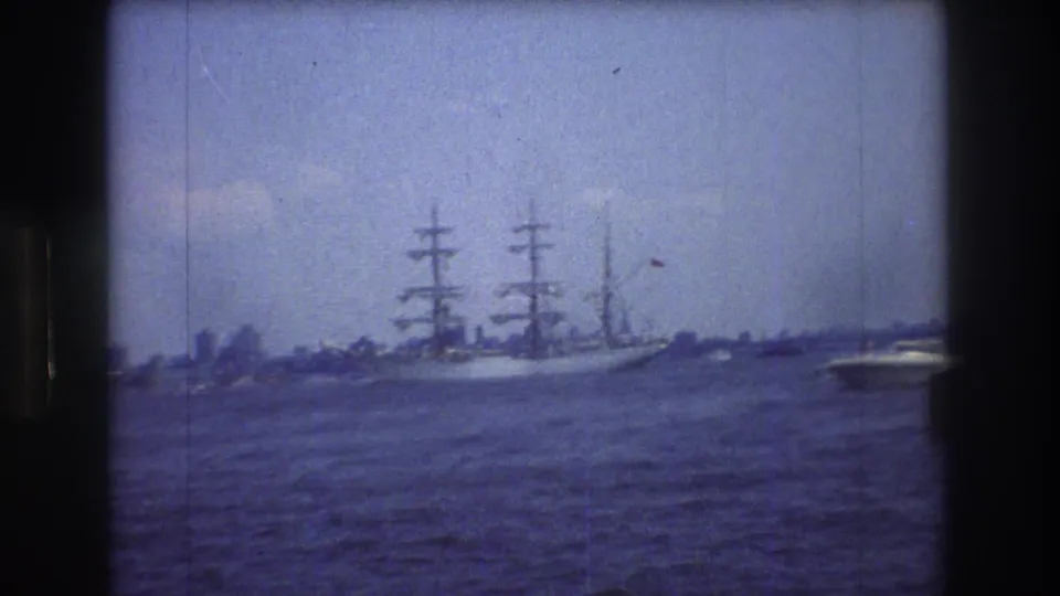 A 1976 8mm view of a multi-masted sailing ship on New York Harbor, seen across choppy water with a distant shoreline and dark frame edges.