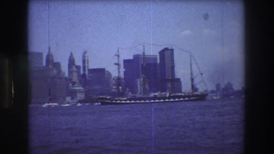 A 1976 home-movie frame of the Manhattan skyline viewed from the harbor, with a multi-masted ship near the waterfront and cranes or tall structures in the background.