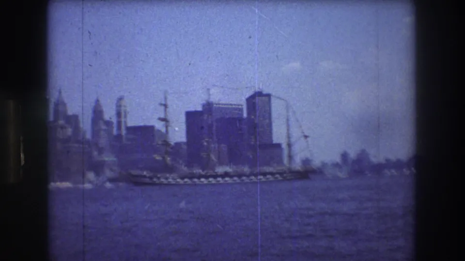 A soft 1976 8mm harbor view of a sailing ship passing before the Manhattan skyline, framed by dark edges and visible small-gauge film texture.
