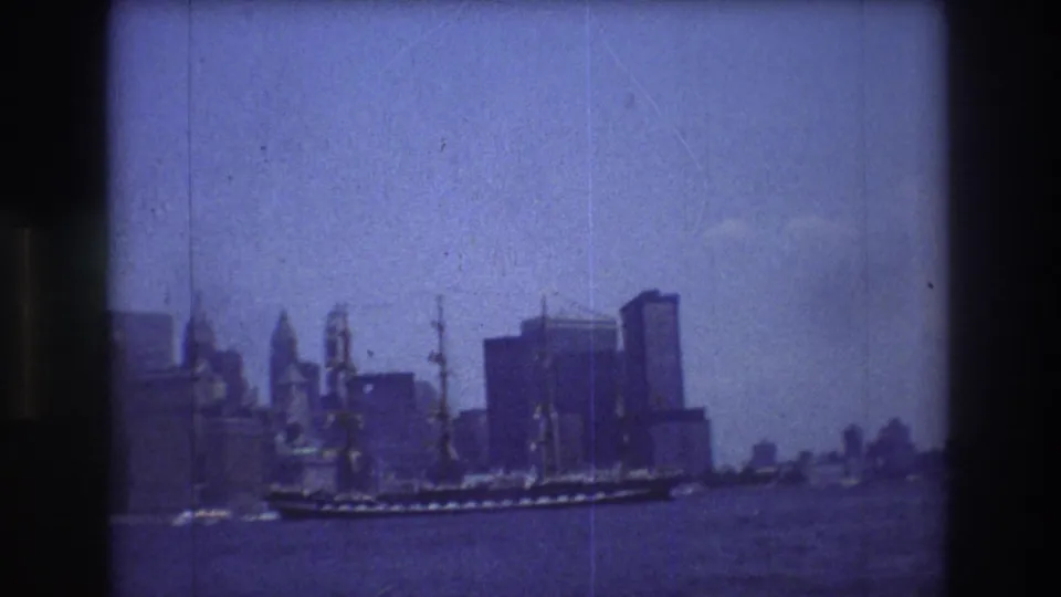 The ship sits low in the frame as it passes the Manhattan skyline, with wide harbor water, skyscrapers, and archival 8mm grain visible.