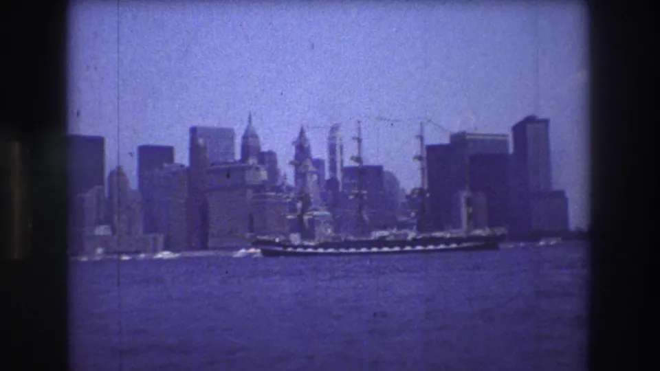 A large sailing vessel continues across the river before the Manhattan skyline, seen through soft, grainy 1976 home-movie color film.