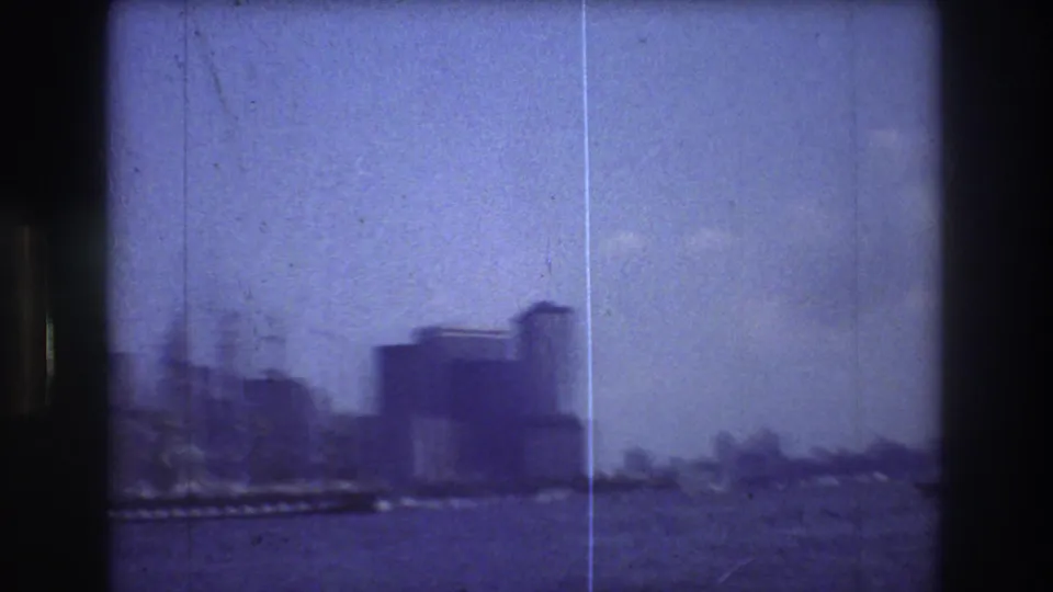 A panning 1976 New York harbor view catches the sailing ship partly at the left edge, with water, skyline blocks, pale sky, and the soft blue-purple cast of the home-movie scan.