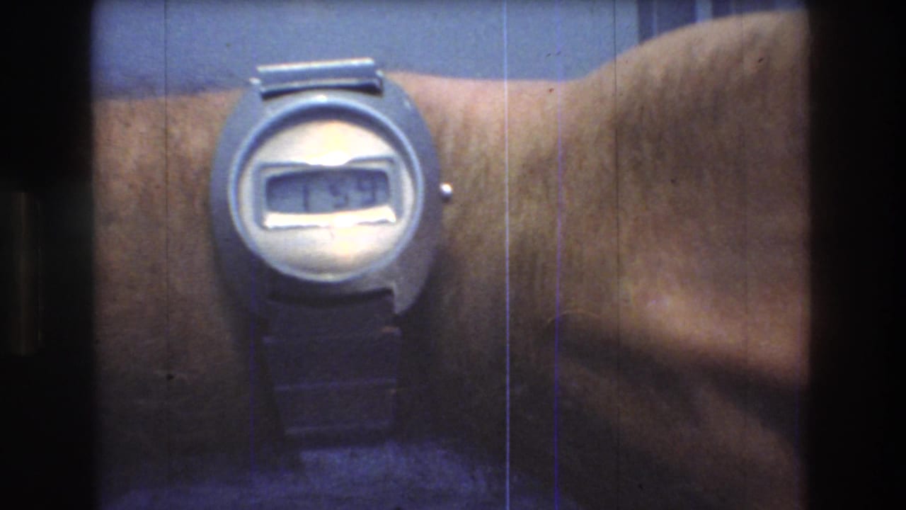 Close-up from 1976 8mm home-movie footage showing a man's wrist with a silver digital wristwatch against a blue background.