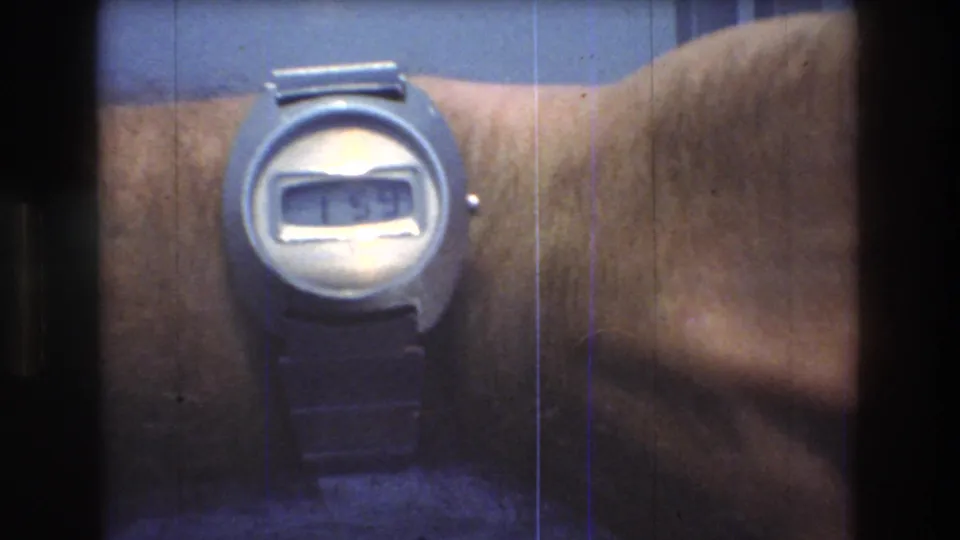 Close-up from 1976 8mm home-movie footage showing a man's wrist with a silver digital wristwatch against a blue background.