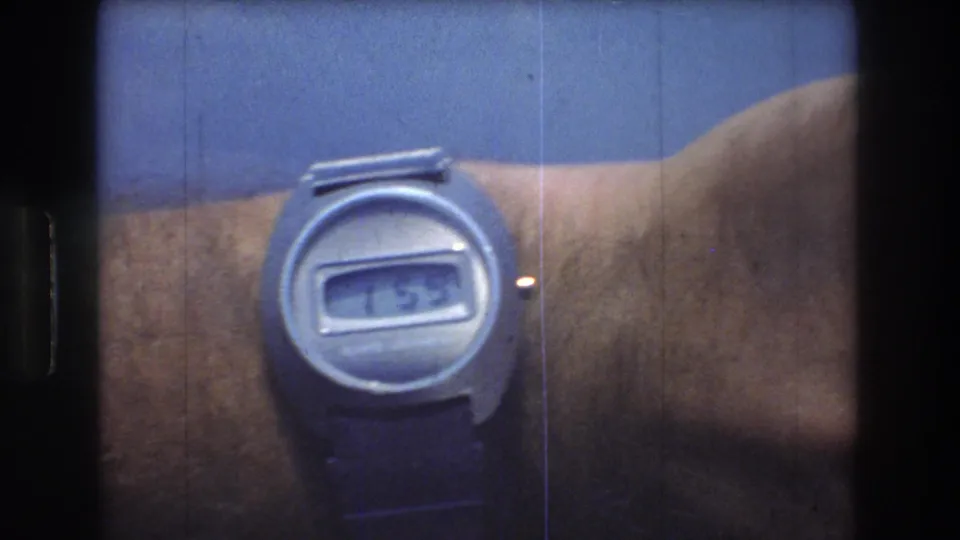 End frame of the 8mm close-up showing a silver digital wristwatch and dark strap on a man's wrist, with film scratches visible over the blue background.