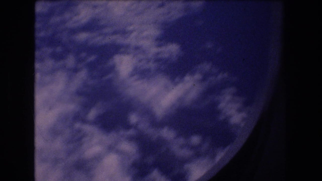 1977 8mm travel footage frame showing white cloud cover and blue sky through a partially visible airplane window, with dark film and window borders around the view.