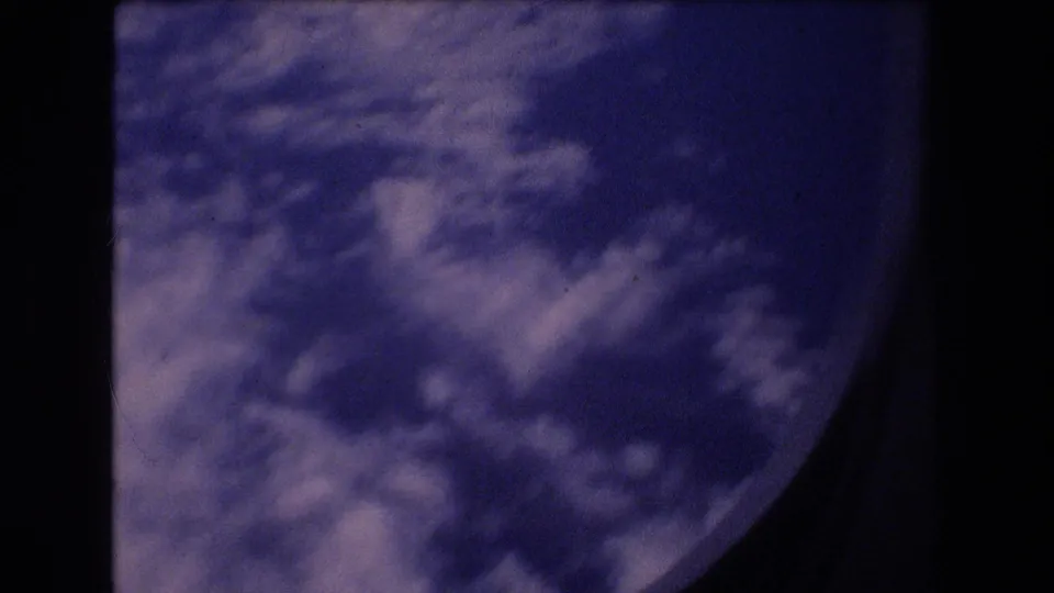 1977 8mm travel footage frame showing white cloud cover and blue sky through a partially visible airplane window, with dark film and window borders around the view.