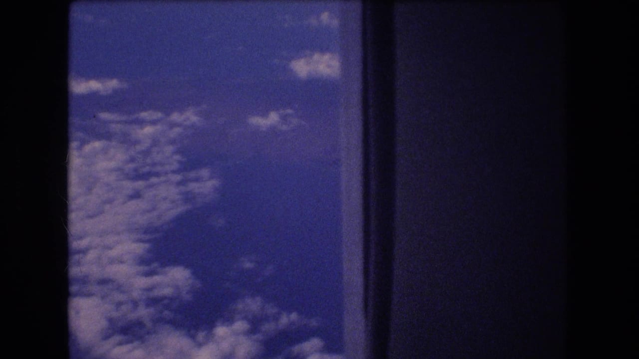 1977 8mm home-movie frame showing blue sky and scattered clouds viewed through a dark airplane window during travel associated by metadata with Honolulu, Hawaii.