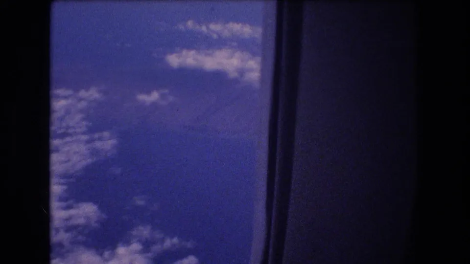 A dark airplane window frame surrounds a blue sky view with scattered clouds in this 1977 8mm travel footage frame.