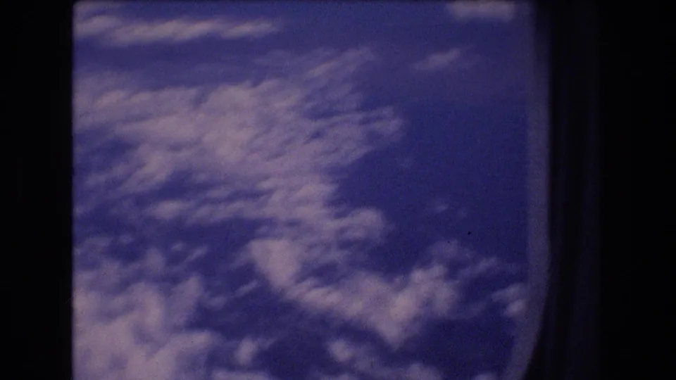Soft blue and white cloud layers fill the lower part of the frame, with the dark airplane window edge still visible in the 8mm scan.