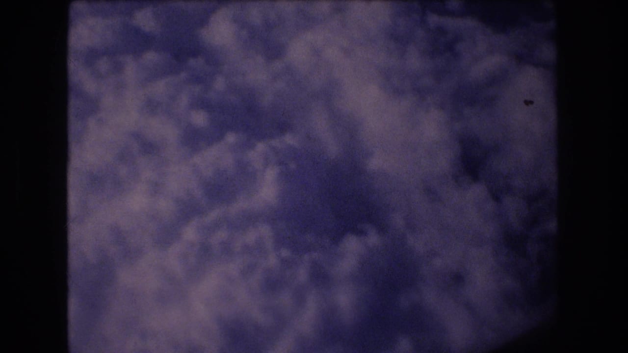 1977 8mm passenger-window view toward dense blue-purple clouds, with the dark aircraft window edge framing the scene.
