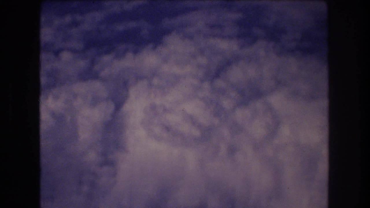 1977 8mm home-movie frame showing layered clouds and blue sky from inside an airplane, with dark window framing along the left and right edges.