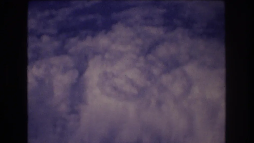 1977 8mm home-movie frame showing layered clouds and blue sky from inside an airplane, with dark window framing along the left and right edges.