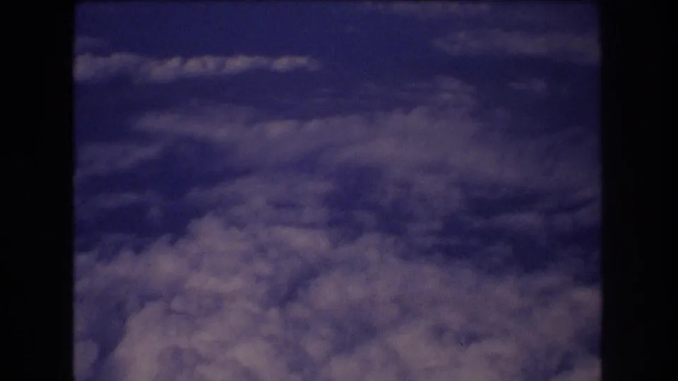 A passenger-window view of cloud tops against deep blue sky, with dark aircraft window structure visible at the side of the 8mm frame.