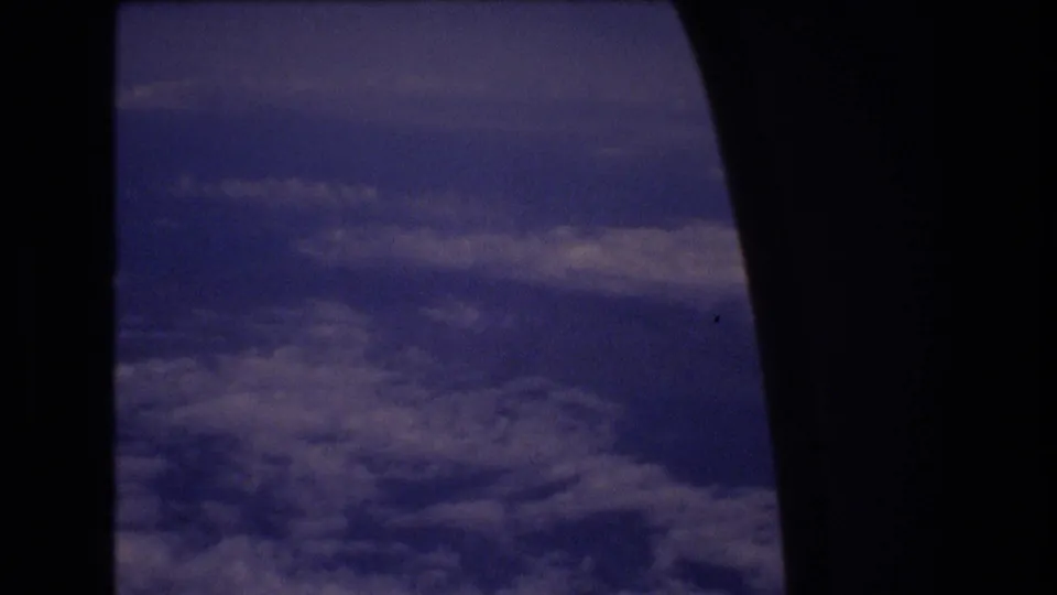 Final 8mm frame looking out from an aircraft toward clouds and blue sky, with a large dark window silhouette along the right side.