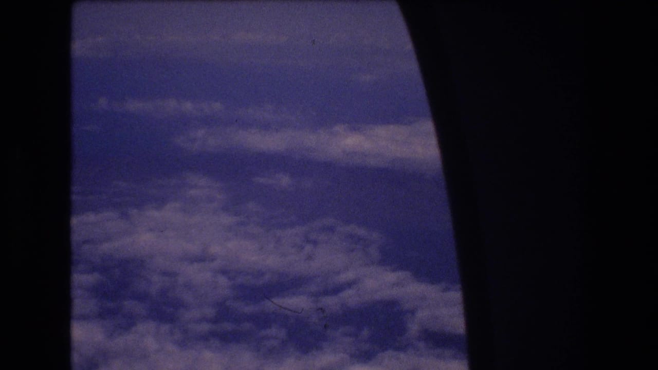 1977 8mm home-movie view from an airplane window, with blue-purple sky and patchy clouds below during a flight associated with Honolulu travel metadata.