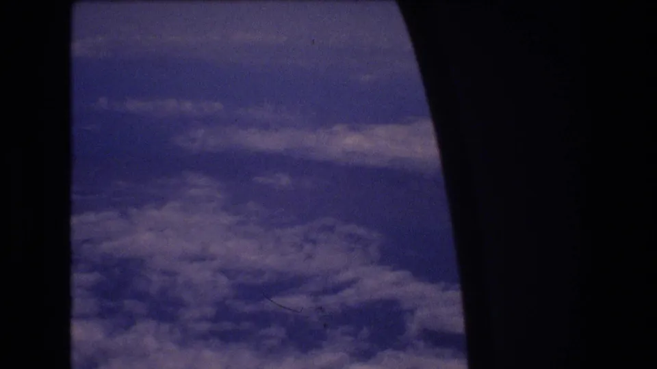 1977 8mm home-movie view from an airplane window, with blue-purple sky and patchy clouds below during a flight associated with Honolulu travel metadata.