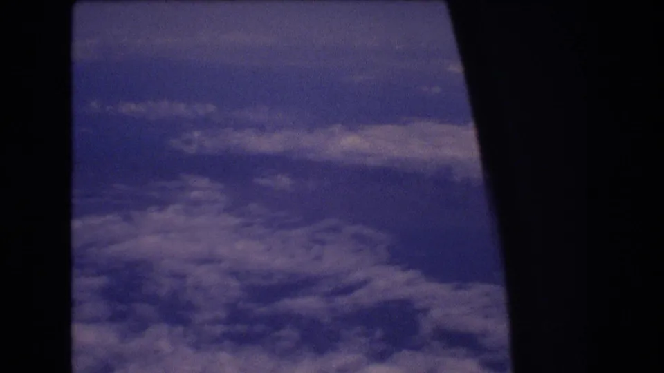 A steady 8mm airplane-window view shows layered clouds beneath a dark window border, with no visible people, aircraft markings, or ground landmarks.