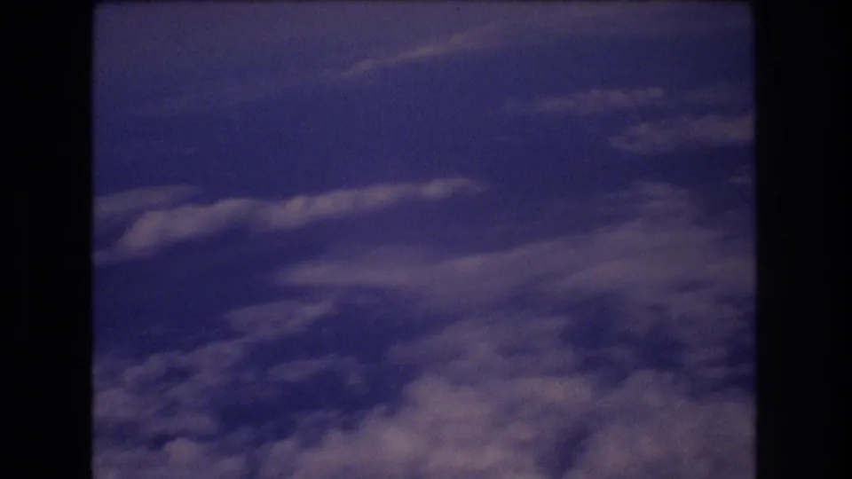 The home-movie frame holds on a blue-violet aerial cloudscape seen through the lower portion of an airplane window during a 1977 travel clip.
