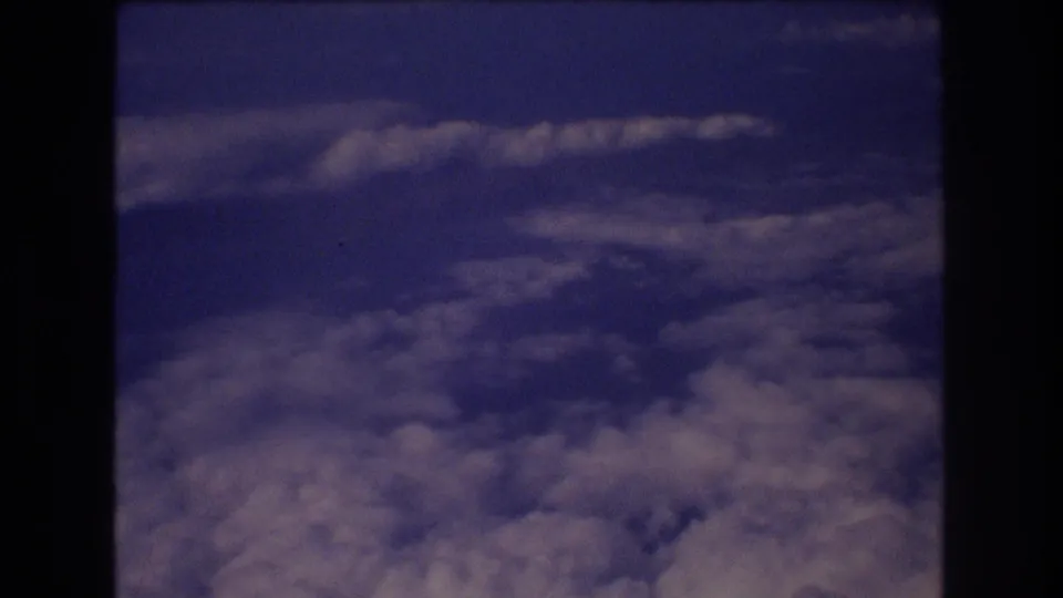 Vintage 8mm travel footage shows a dense cloud layer below and deep blue sky above, framed by dark airplane-window edges during a Honolulu-associated flight.
