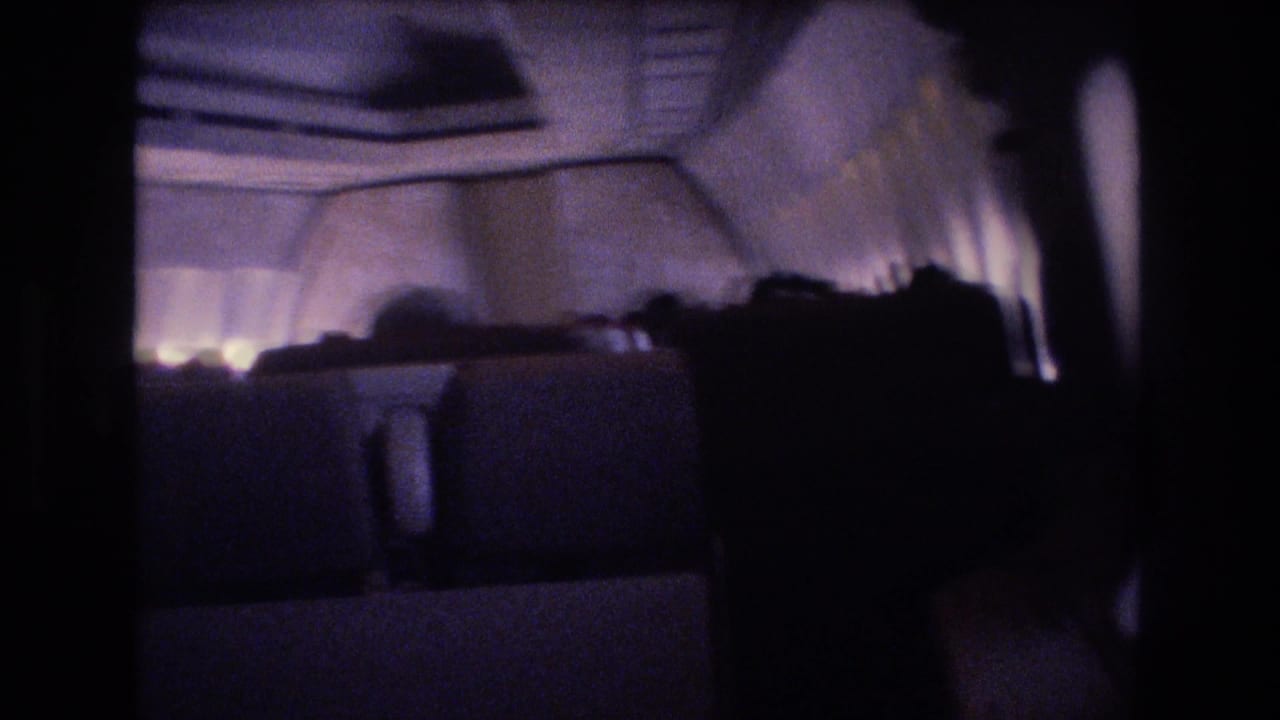 1977 8mm home-movie frame from a dark airplane cabin, with passengers seated in silhouette beneath dim cabin lighting and small illuminated windows.