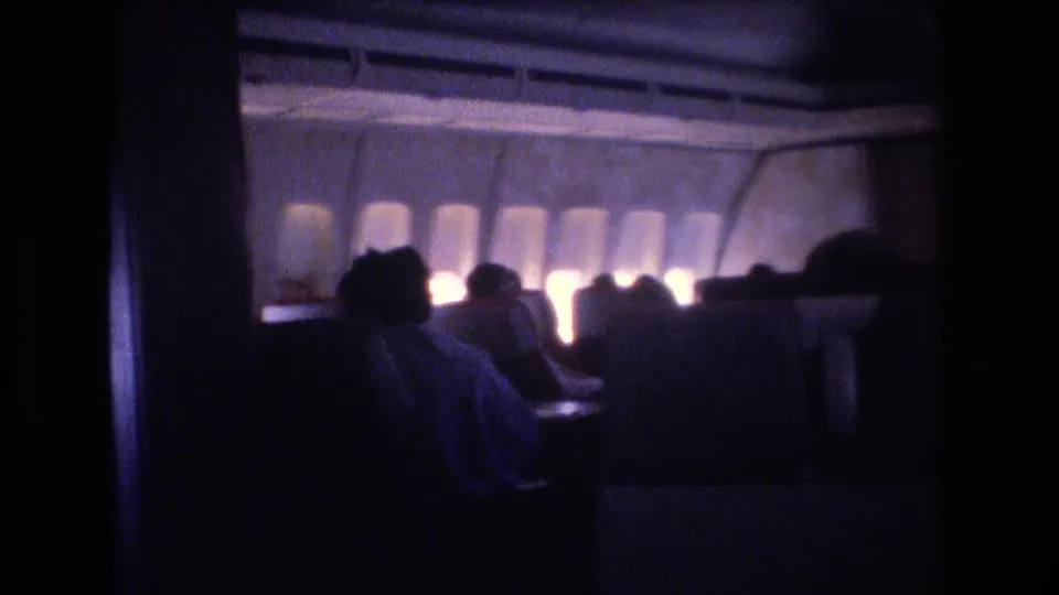 Dark 8mm cabin view with seated passengers in silhouette, rows of airplane windows glowing faintly, and seat backs filling the foreground.
