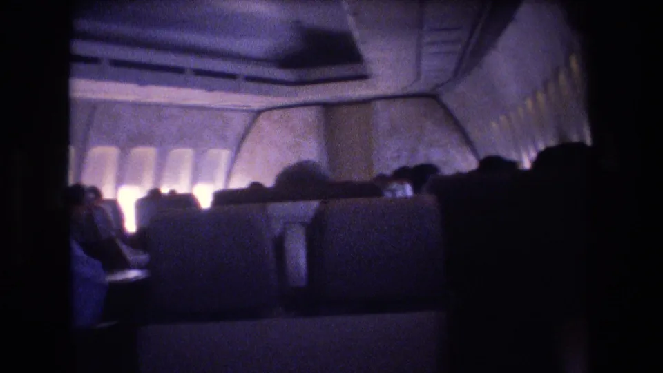 Handheld 8mm view across a dark airplane cabin, showing rows of seats, glowing window openings, overhead panels, and passengers mostly reduced to silhouettes.