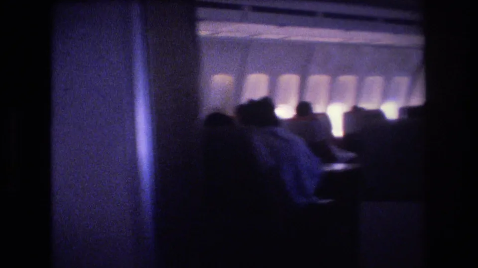 Dim 1977 home-movie frame inside an airplane cabin, with a dark vertical partition or aisle edge, seated passengers, and softly glowing windows in the background.
