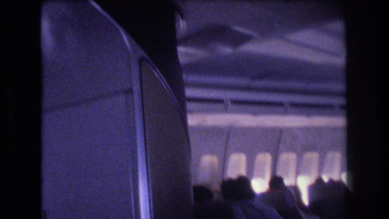 1977 8mm home-movie frame inside a commercial airplane cabin, with first-class-style seat backs in the foreground and passengers silhouetted against bright window light.