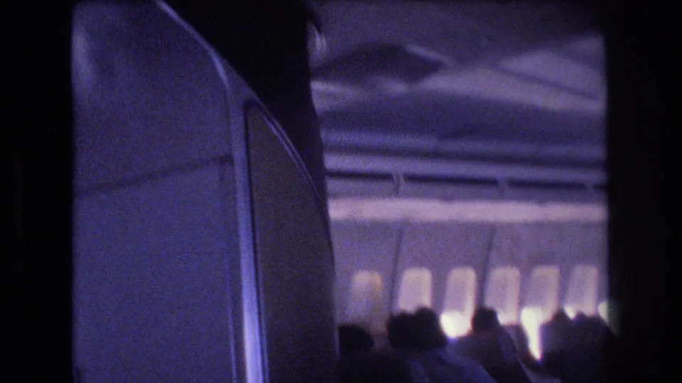 1977 8mm home-movie frame inside a commercial airplane cabin, with first-class-style seat backs in the foreground and passengers silhouetted against bright window light.