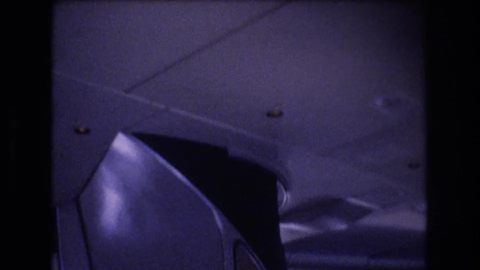 Soft 8mm view of a commercial airplane interior, showing ceiling panels, overhead storage, and a foreground seat back in a blue-purple low-light cast.