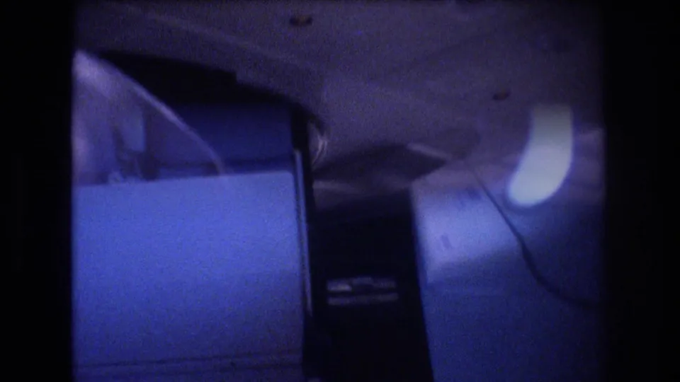 Close 8mm cabin frame showing a large airplane seat back, dark aisle area, and a bright oval window or cabin light shape in the background.
