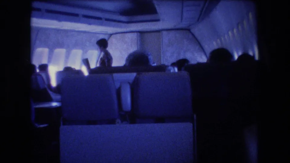 1977 8mm airplane-cabin frame with a flight attendant standing near the aisle and passengers seated in silhouette along the window row.