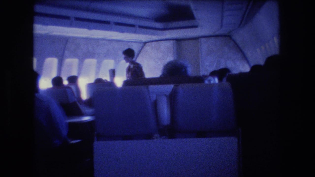 1977 8mm home-movie frame showing a blue-tinted airplane cabin interior with seated passengers, cabin windows, overhead panels, and a flight attendant standing near the aisle.