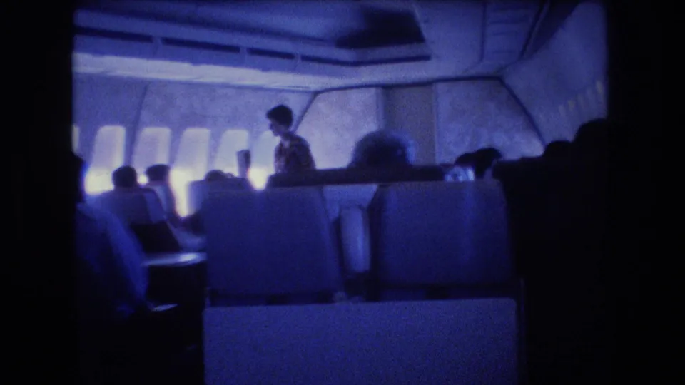 1977 8mm home-movie frame showing a blue-tinted airplane cabin interior with seated passengers, cabin windows, overhead panels, and a flight attendant standing near the aisle.