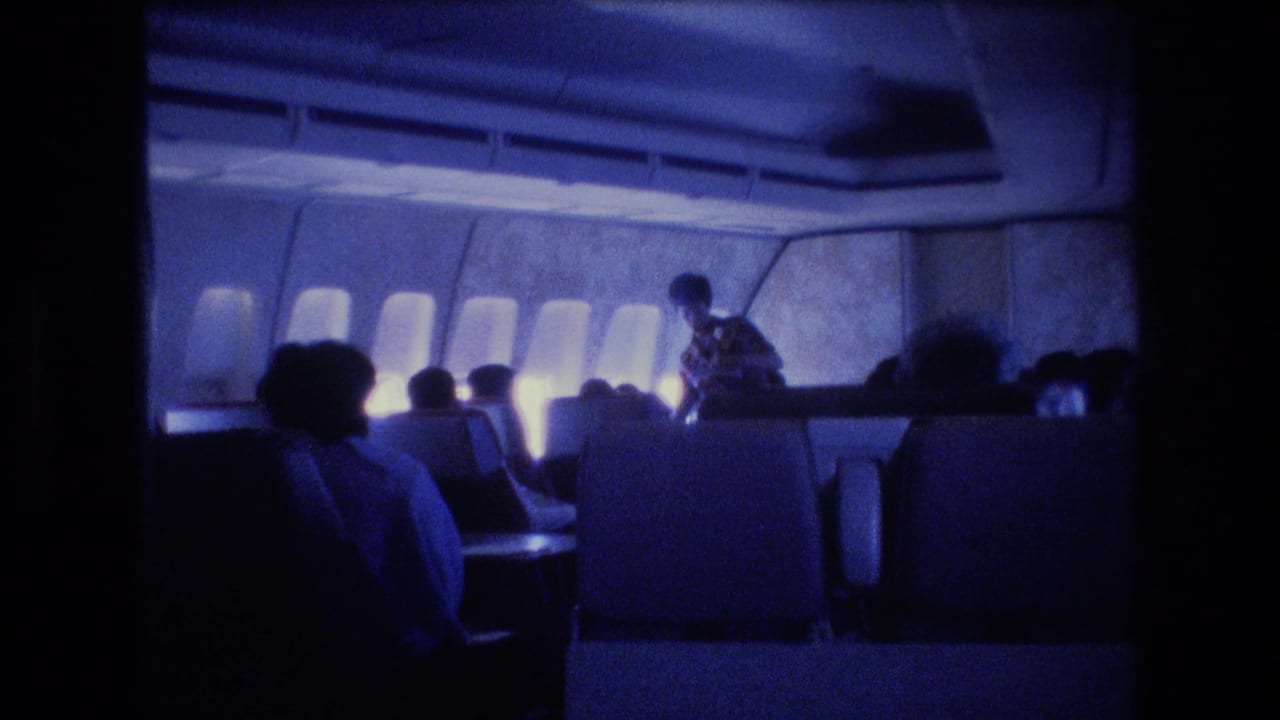 Blue-tinted airplane cabin with passengers seated and a flight attendant moving in the aisle.