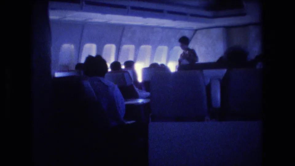 1977 home-movie frame showing seated passengers in a blue-tinted airplane cabin, with window light behind them and a flight attendant partly visible near the right side.