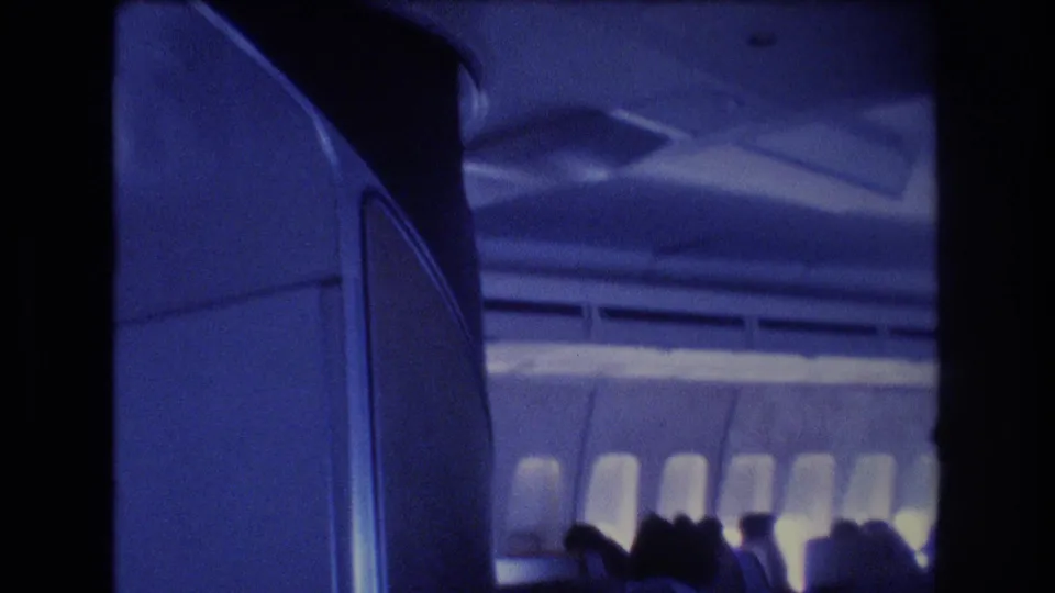 1977 8mm frame with a closer airplane-cabin view, showing curved interior paneling, overhead fixtures, bright windows, and passengers seated along the side.