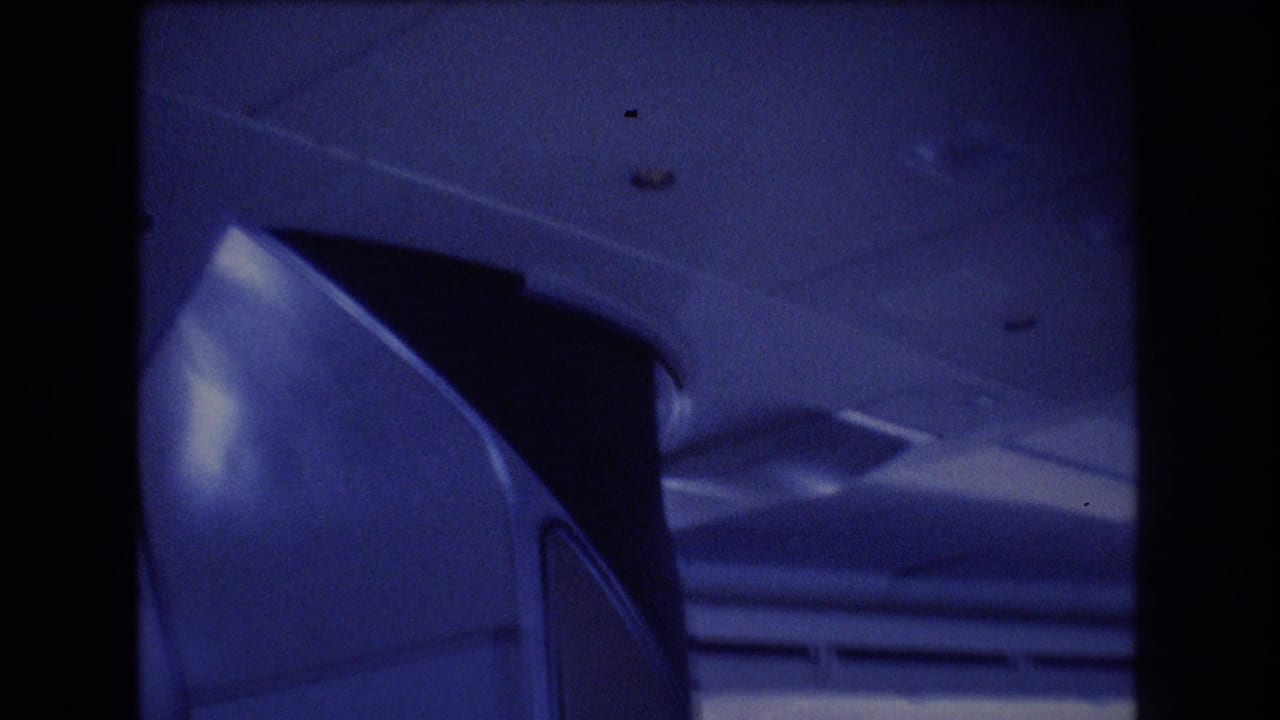 Blue-tinted 1977 8mm frame showing part of an airplane cabin interior, including overhead fixtures and the curved side of a compartment or seat area.