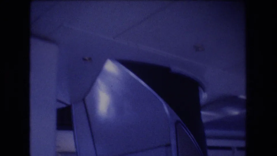 1977 home-movie frame from inside an airplane cabin, with overhead paneling, ceiling details, and a curved interior structure visible in soft blue light.