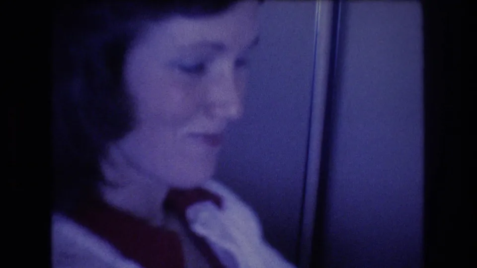 1977 8mm frame showing a uniformed adult woman, identified by metadata as a flight attendant, inside an airplane cabin with blue-tinted light and soft focus.