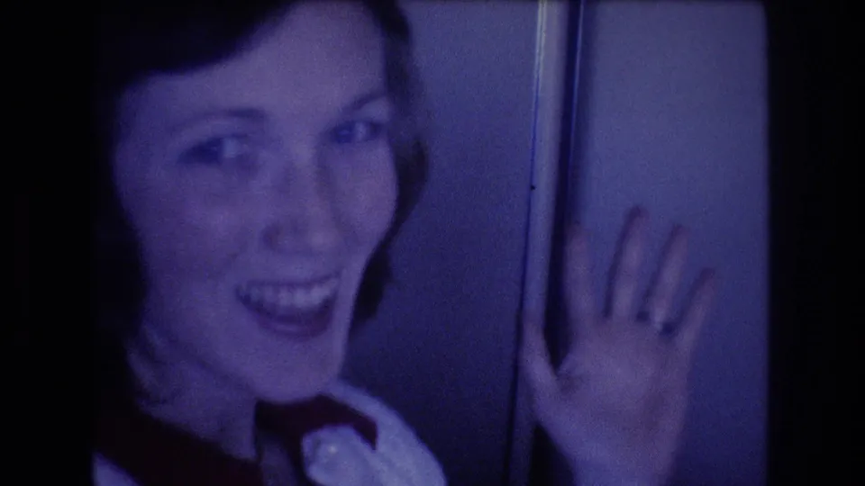 A young woman in a white collared shirt and dark tie smiles and raises her hand toward the camera during a 1977 home-movie airplane shot associated with Honolulu travel metadata.