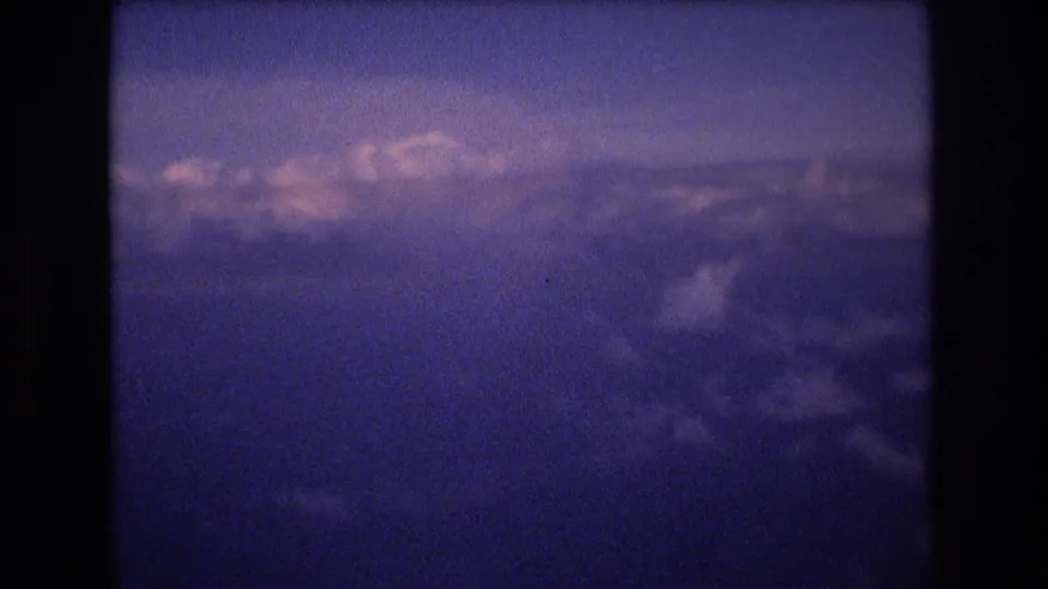 The camera looks out over textured cloud banks under a hazy sky, continuing the 1977 8mm airplane travel view supplied in the clip metadata.