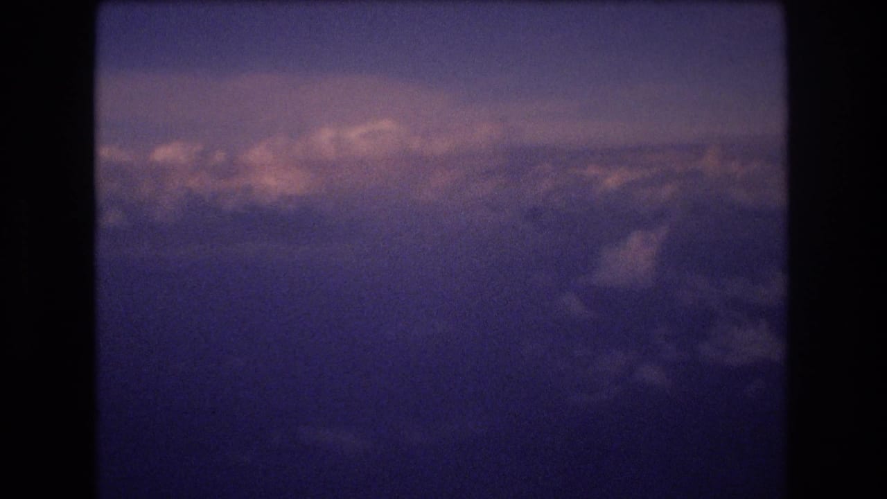 1977 8mm home-movie frame showing an aerial view of cloud layers with purple-blue shadows and pink light near the horizon, associated by metadata with Honolulu travel.