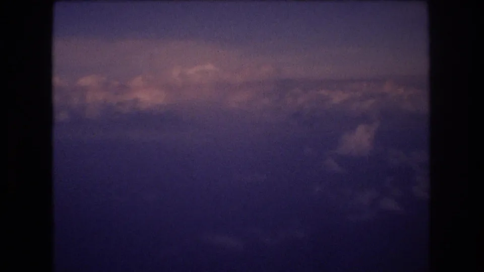 Small-gauge film frame showing layered clouds below a hazy purple-blue sky, with the home-movie scan retaining grain and soft exposure.