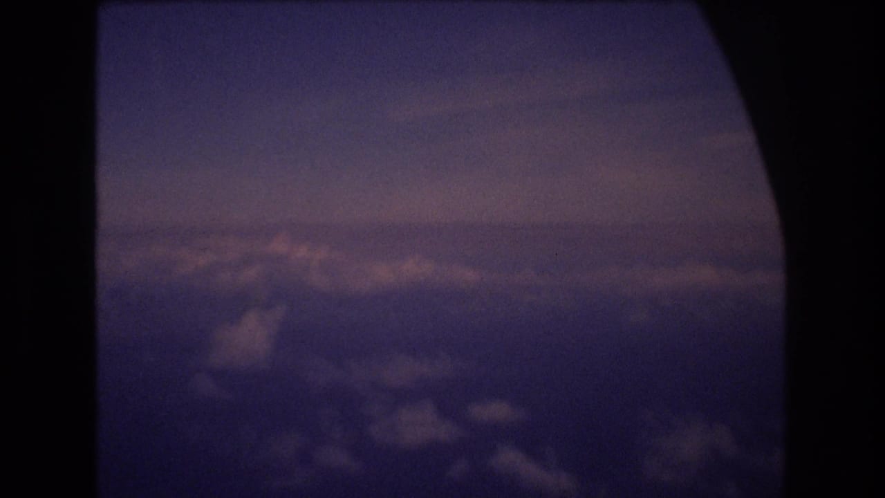1977 8mm home-movie frame showing clouds and twilight sky seen through a dark airplane window during travel associated with Honolulu, Hawaii metadata.